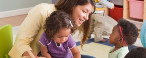 two children in a child care setting with an adult sitting with them. The adult woman is smiling and looking at one of the children.