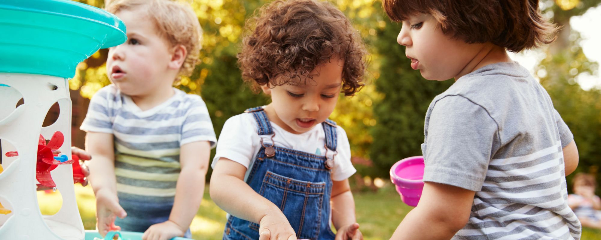 photo of three toddlers, one with blonde hair and two with dark hair. they are playing at a water table