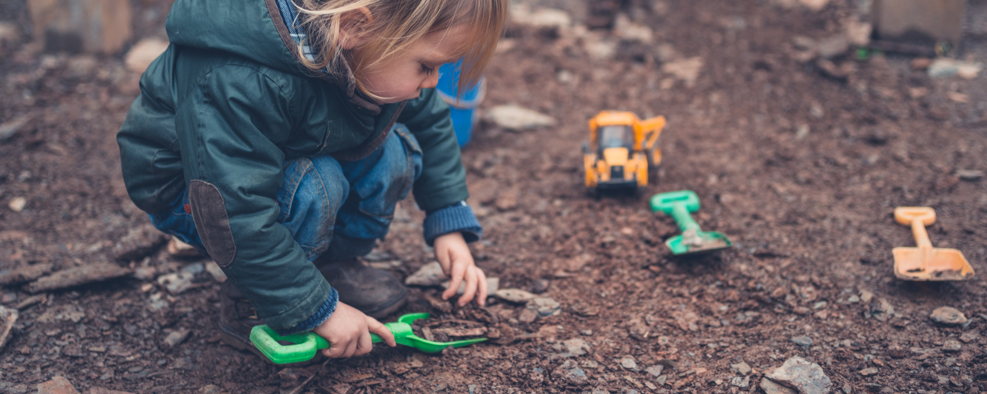 preschooler crouched down playing in the dirt with a shovel. There are other shovels and tools and toys around them. They are very interested in their surroundings.