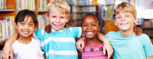 four kids, around age 4 are standing together in a library. There are book shelves in the background. The kids have their arms around each other. They are from different backgrounds, wearing colourful clothing and all smiling happily.