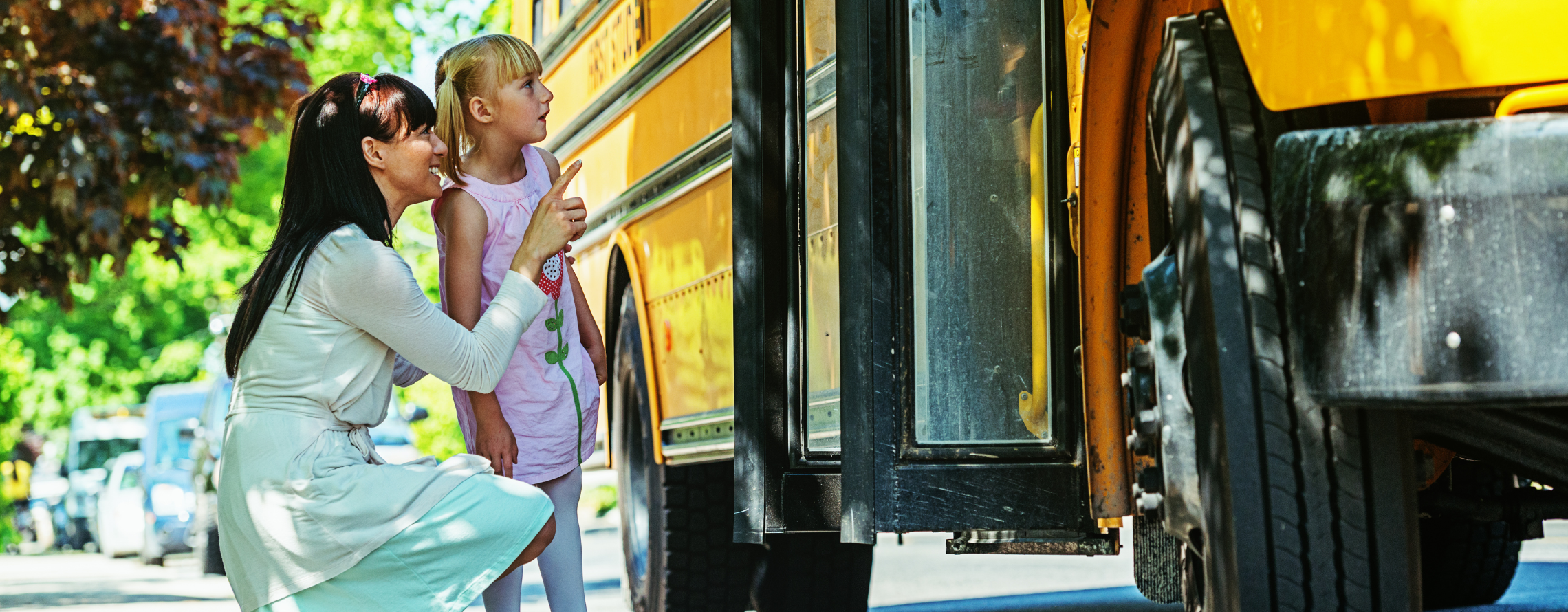 a little girl with her caregiver standing next to a school bus. Its implied that it's the first day of school.