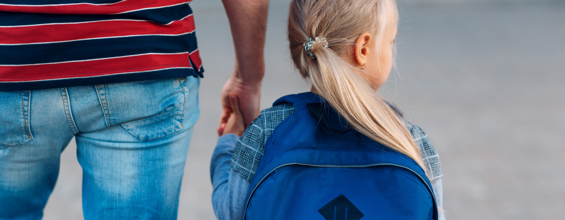 little blonde girl with a pony tail holding a parent's hand. she has a backpack on and the vibe of the photo is that she's nervous on her first day of school.
